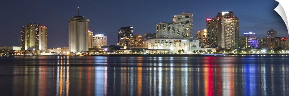 Panoramic photograph of the New Orleans skyline lit up at night and reflecting colorful bands onto the Mississippi River.