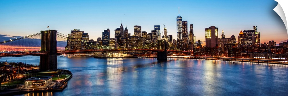 Panoramic view of the New York City skyline illuminated at night, with the Brooklyn Bridge, from across the water.