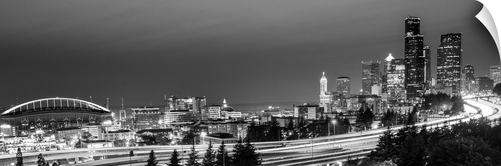 Panoramic photograph of the Seattle skyline with the stadium on the left and light trails from traffic on the highway.