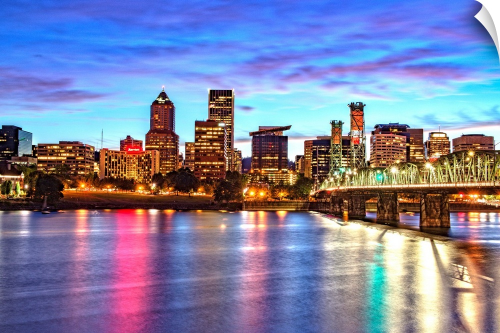 Photograph of the Portland, Oregon skyline lit up at sunset with colors reflecting onto the Willamette River in the foregr...