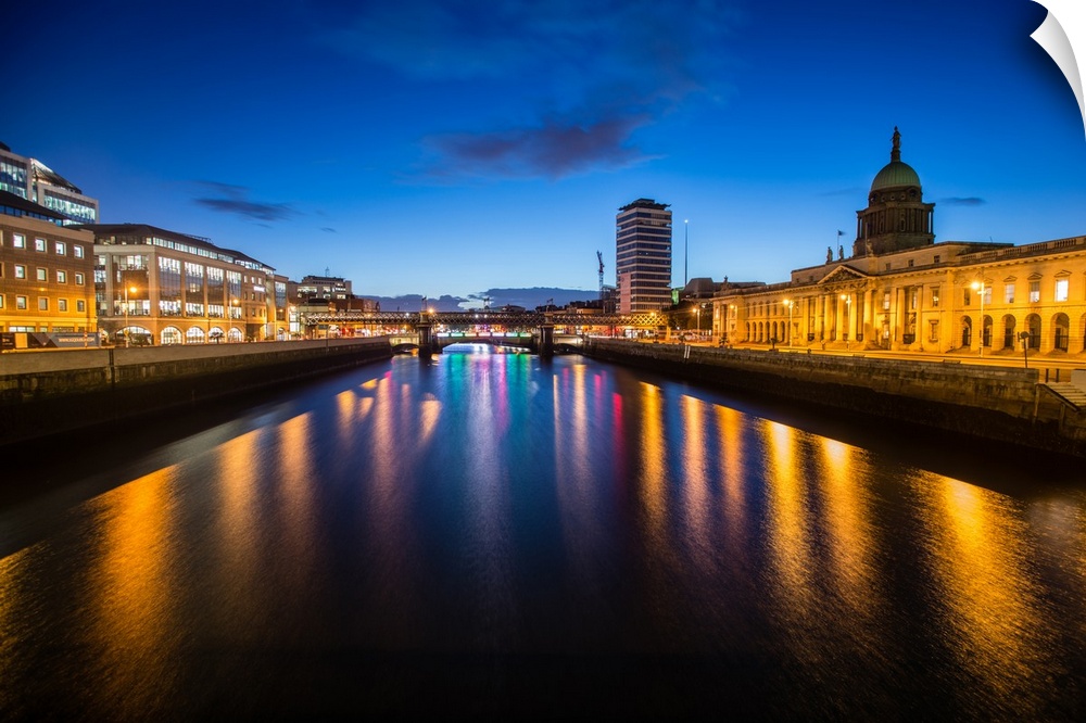 Photograph going straight down the River Liffey with buildings, highlighting The Custom House, on the sides lit up at night.
