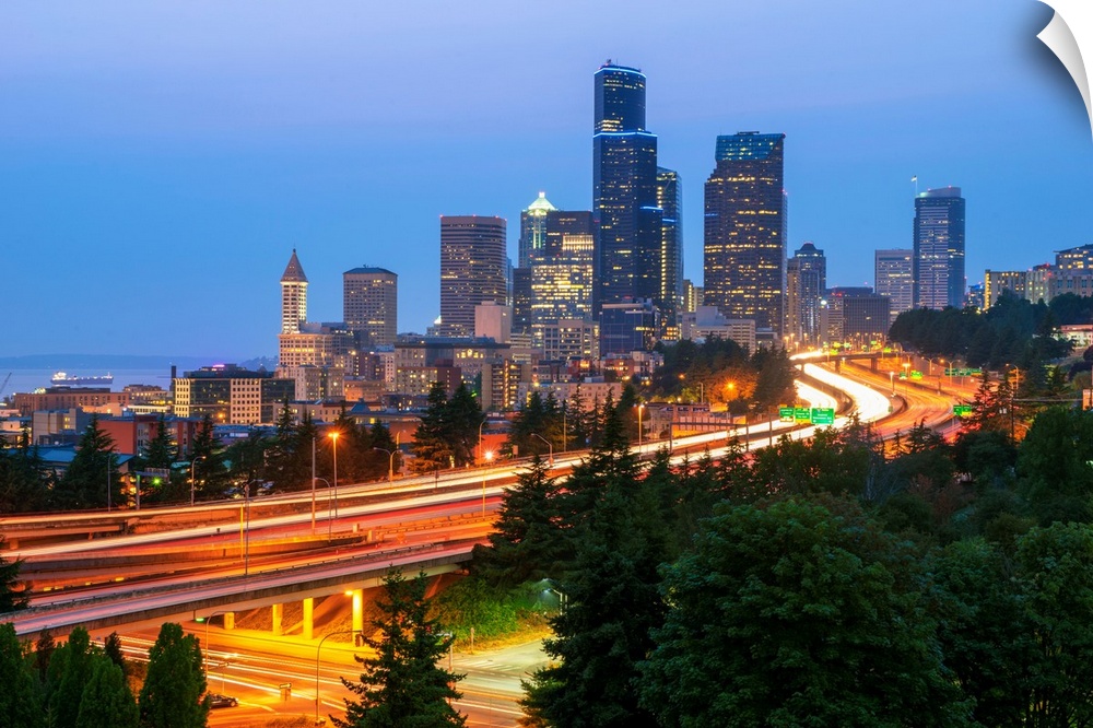Photograph of the Seattle skyline at dusk with light trails from the cars on the highway in the foreground.