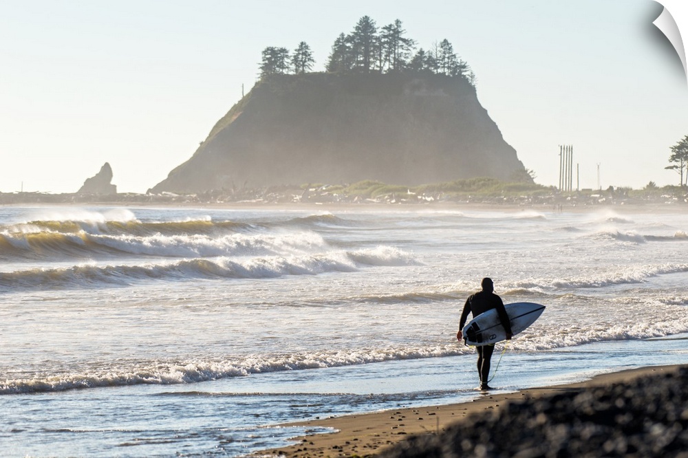 Photograph of a surfer walking on the shore of La Push Beach in Washington, with misty cliffs in the background.