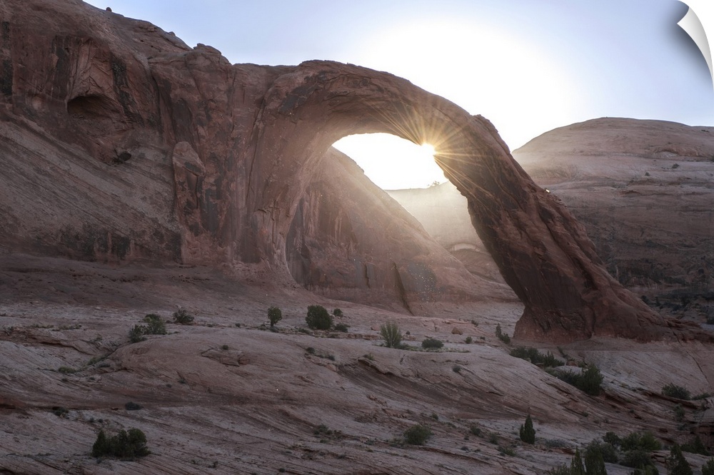 The sun peeking behind the Corona Arch with a view of the desert landscape in Arches National Park, Utah.