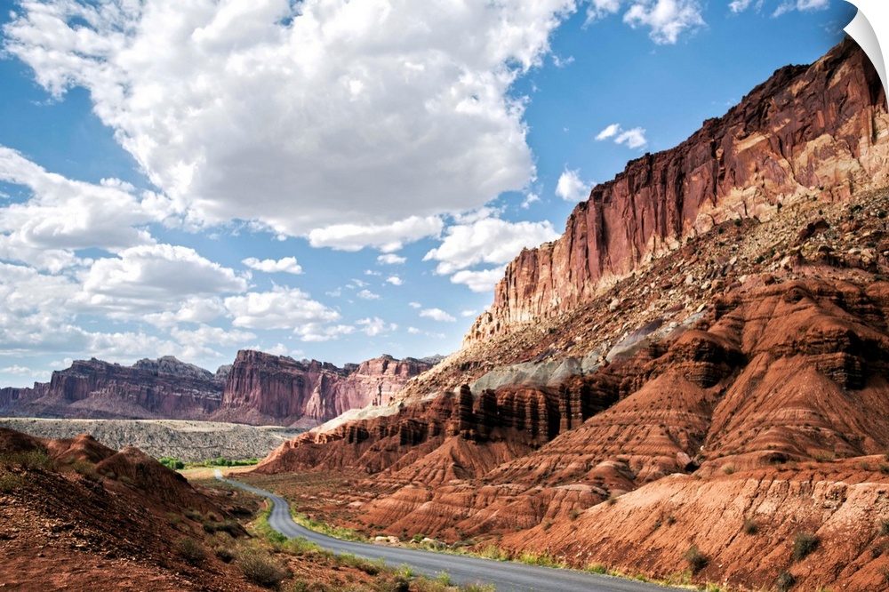 A road winds through Capitol Reef National Park while puffy clouds scatter across a bright blue sky.