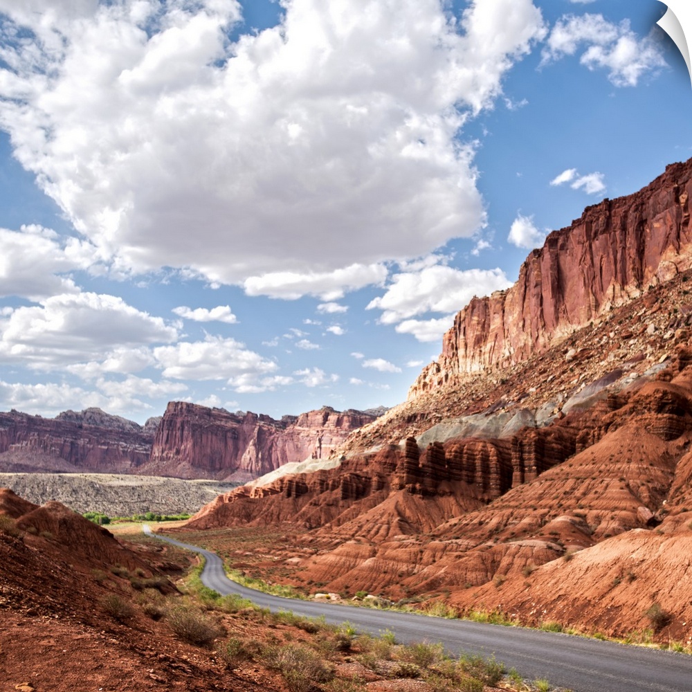 A road winds through Capitol Reef National Park while puffy clouds scatter across a bright blue sky.