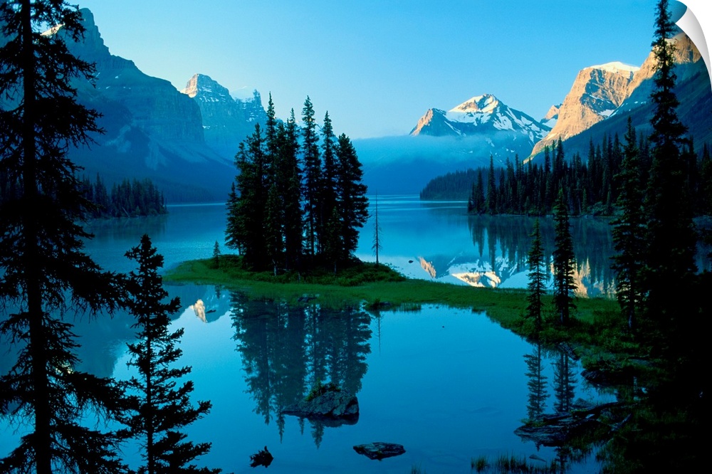 The northern landscape of evergreen trees and mountains reflect on the still waters of a lake in the morning.