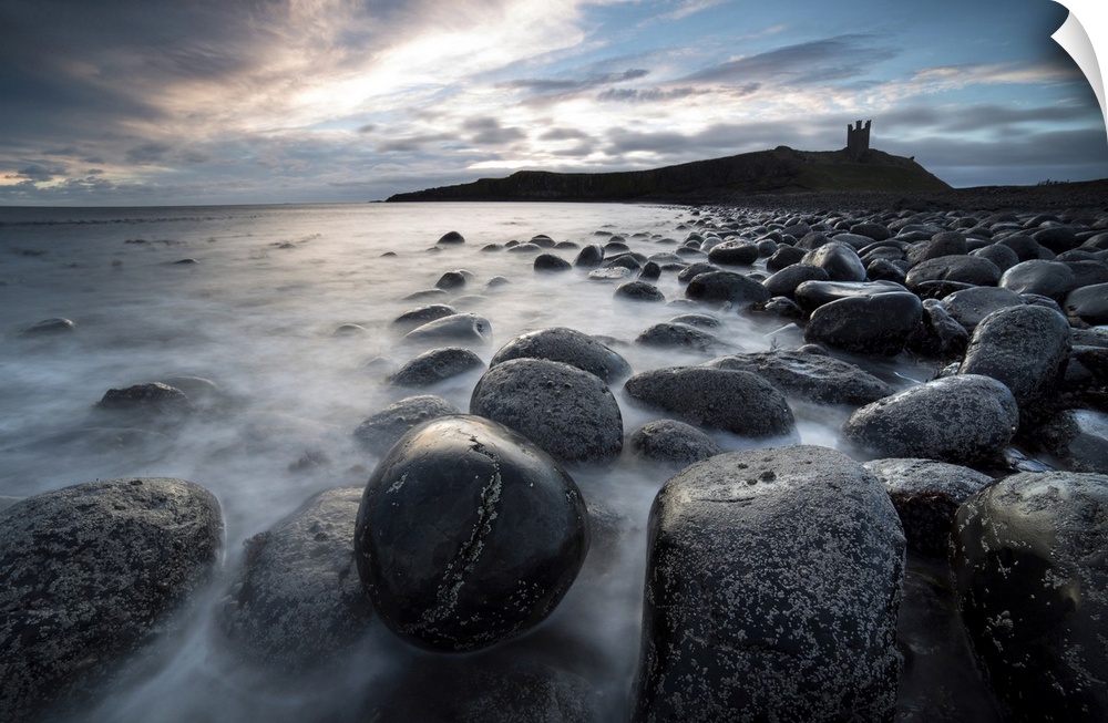 View at sunrise towards the ruin of Dunstanburgh Castle from the beach of basalt boulders known as The Rumble Churn at the...