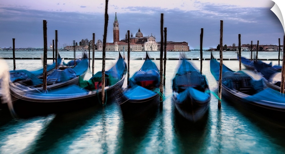 Big photograph of empty gondolas tied to poles in Venice, Italy with buildings in the background.