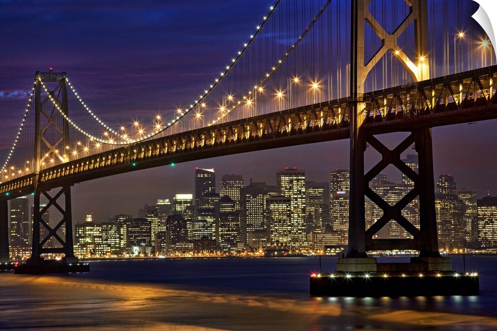 The Bay Bridge is shining brightly under a night sky with the San Francisco skyline shown in the background.