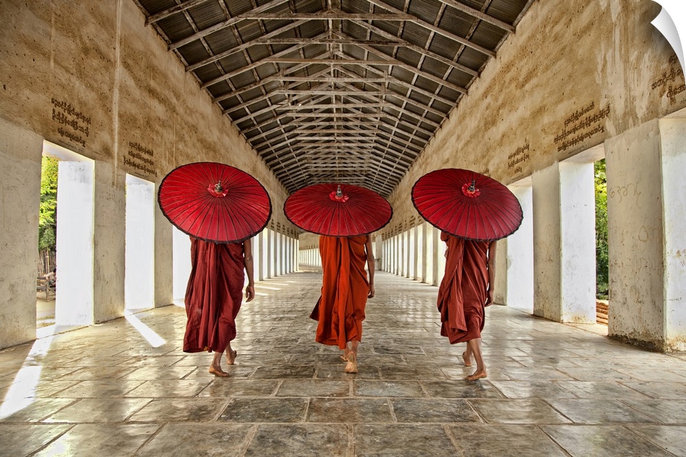 Three monks in bright red walking in a row with umbrellas down a neutral open hallway.