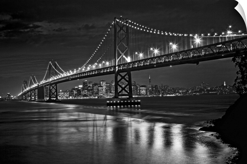 This landscape is a high contrast, monochromatic photograph that shows a bridge in the fore ground and glowing city skylin...