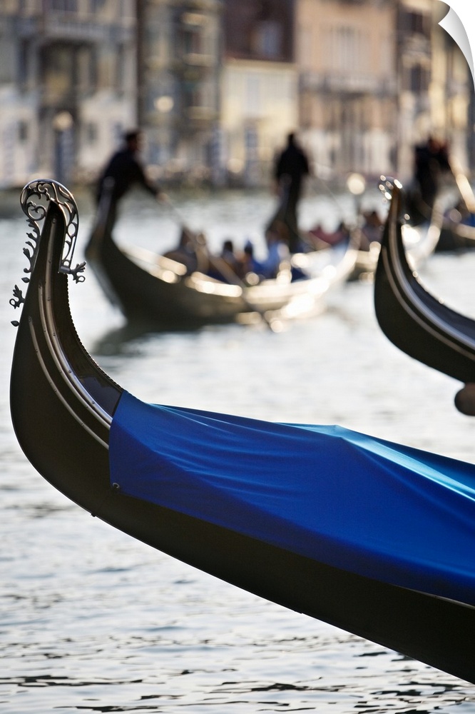 Vertical photograph of the front tip of a Venetian gondola hanging over the water.  In the background is a softly blurred ...