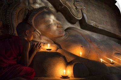 Young Burmese monk praying by candlelight by reclining Buddha