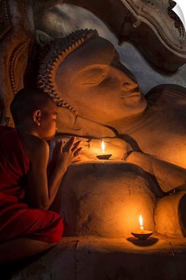 Young Burmese monk praying by candlelight by reclining Buddha