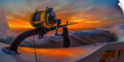 The reel of a big game fishing rod on the side of a boat, with the setting sun behind