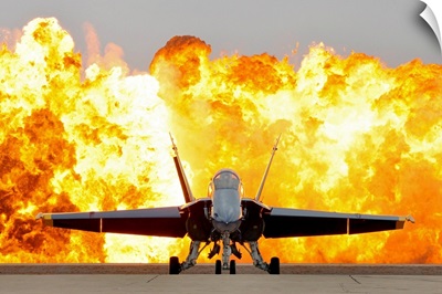 An F/A 18 Hornet sits on the flight line as a wall of fire detonates behind it