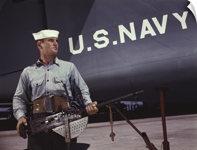 Sailor holding a .30-caliber machine gun in front of a U.S. Navy plane at Naval Air Base