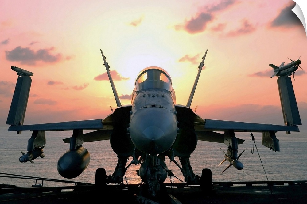 Military photograph of a jet aircraft on the deck of a carrier ship, with wings folded up and the sun rising behind the co...