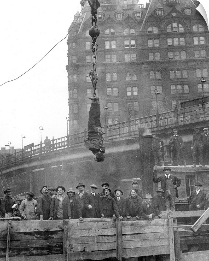 American magician. Houdini in a straitjacket suspended in midair over a subway excavation in New York City; photographed i...