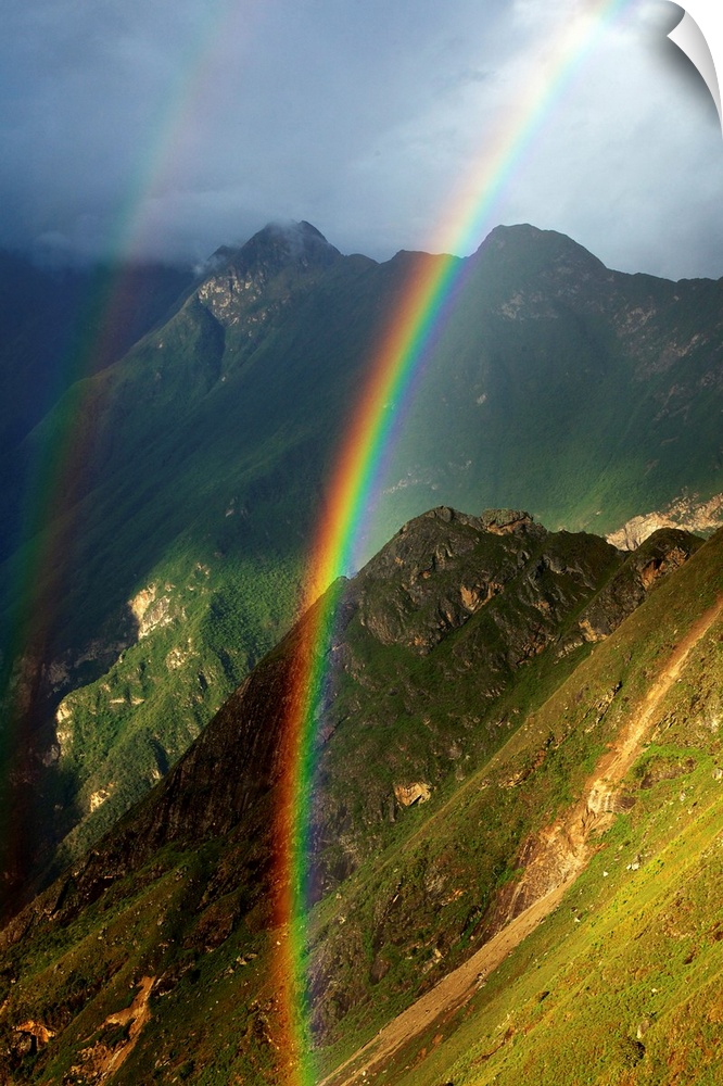 Vertical photograph of a double rainbow over green mountains on a cloudy day.