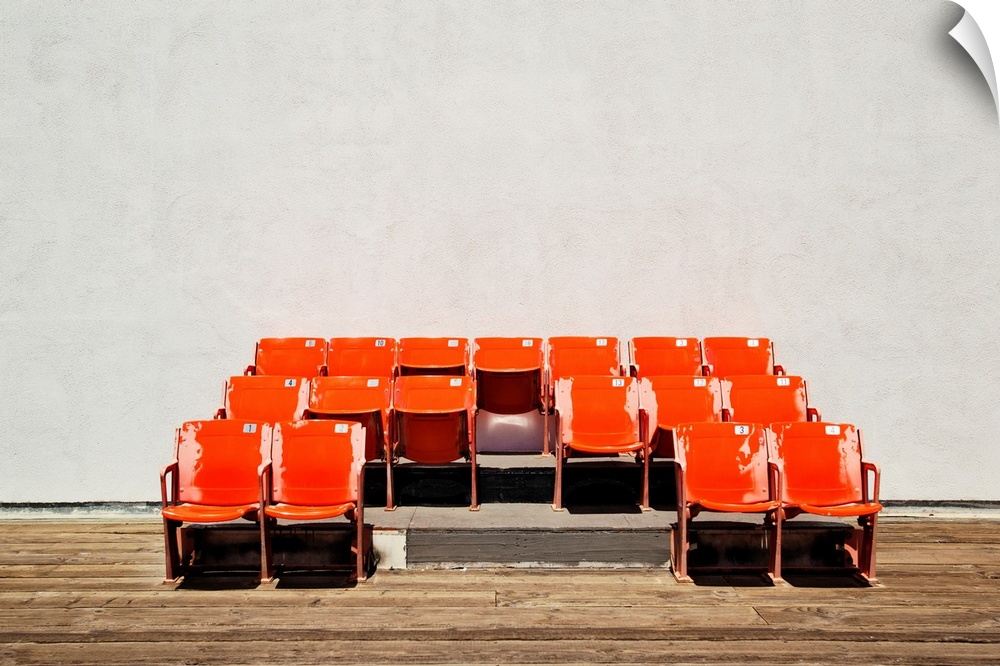 Photograph of three rows of bright orange theater seats against a plain wall and wooden floor.