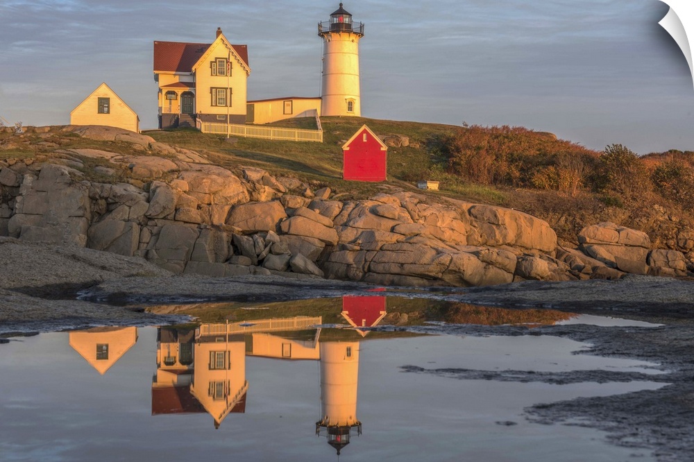 Nubble Lighthouse on a rocky cliff at sunset in Cape Neddick, Maine, reflected in the ocean water below.