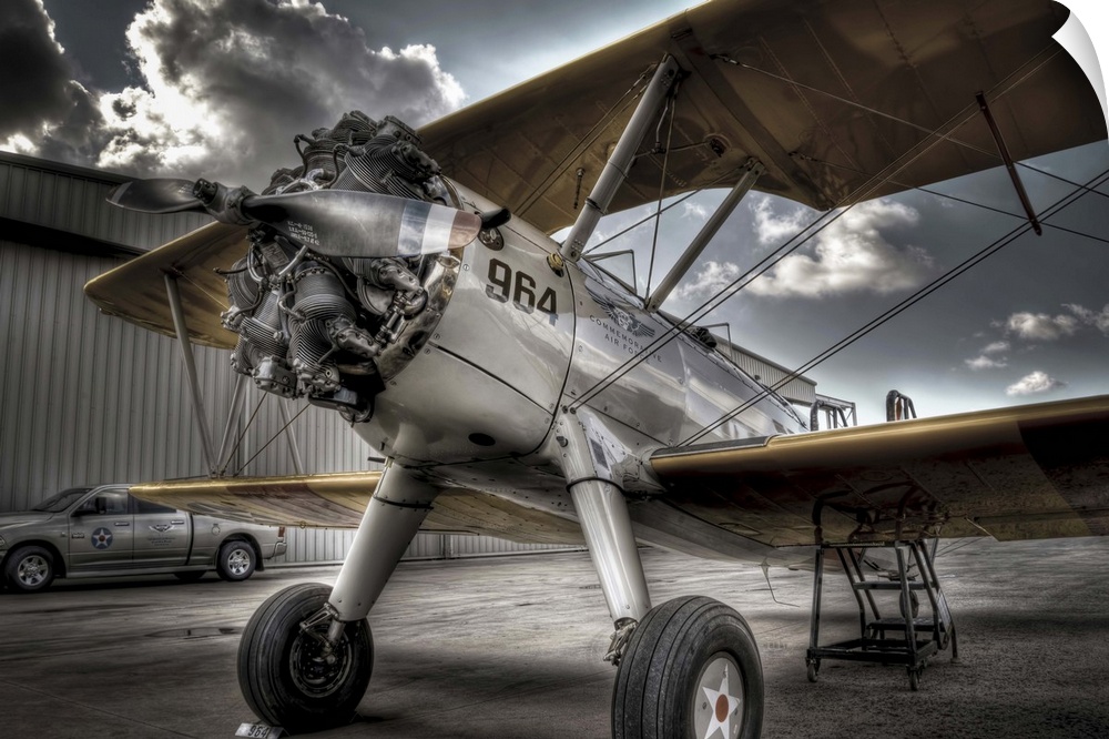 A HDR photograph of a vintage air craft parked on the tarmac outside a hanger and a sky full of large cumulous clouds. The...