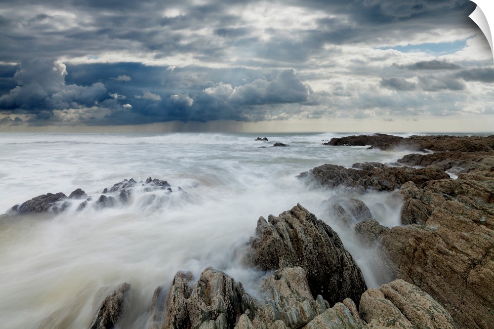 Jagged rocks on coastline with white surf under grey storm clouds