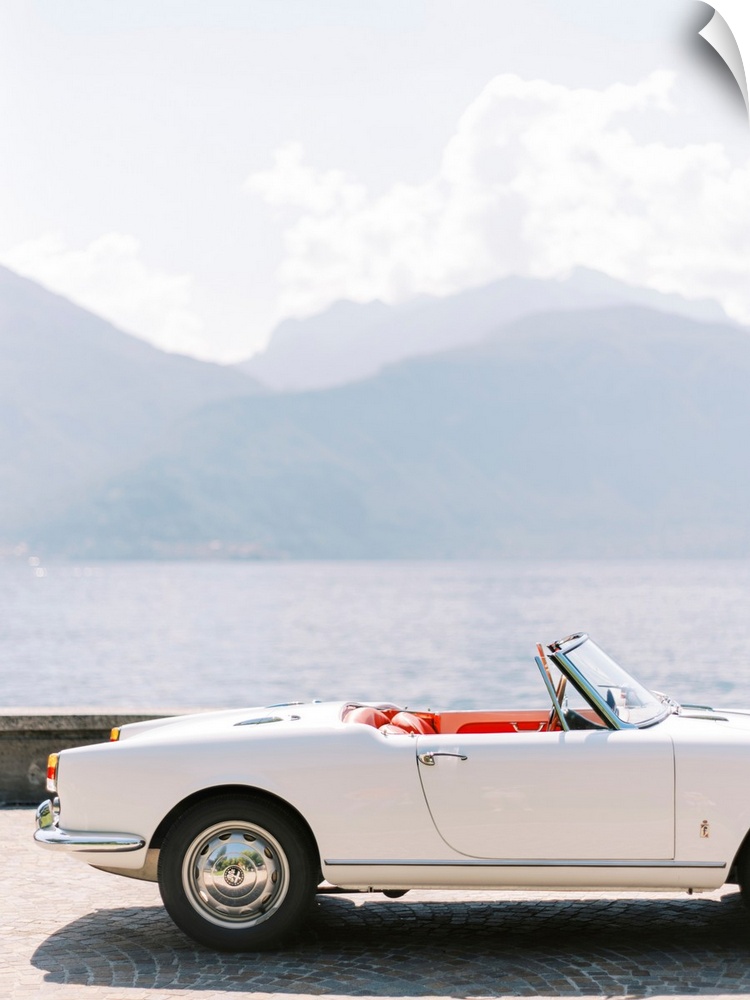 A photograph of the exterior of a white antique convertible sports car with red leather interior, parked in front of Lake ...