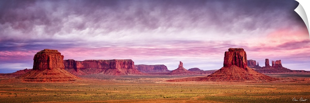 Panoramic photograph of Monument Valley with a pink and purple sunrise above.