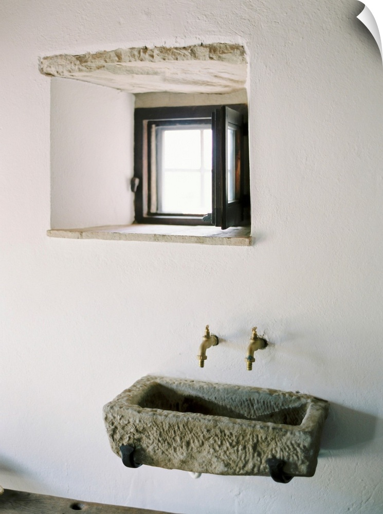 A photograph of a rustic stone wall basin underneath a small window in a white stucco mediterranean dwelling.