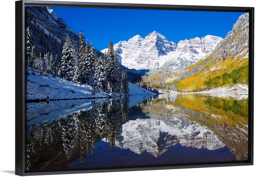 A snow covered mountain range with tall trees in front reflects beautifully into a body of water.