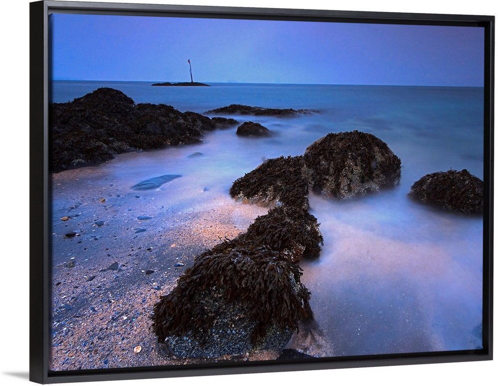 The landscape wall art is a time lapse photograph of waves washing on shore between kelp covered rocks.