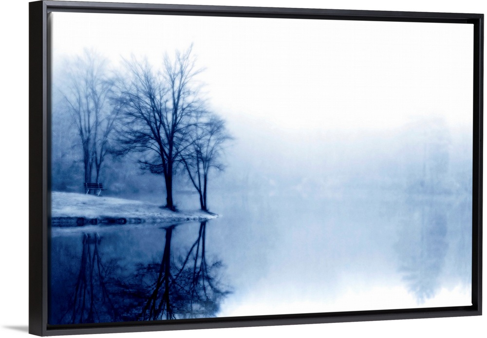 A photograph taken of a lake with bare trees off to the left side and more trees behind dense fog toward the back of the l...