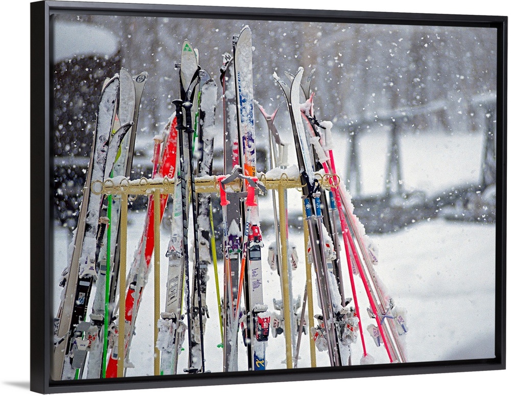 In this photograph multiple skis are stored on an outside rack as large white snowflakes fall all around. Blurred backgrou...