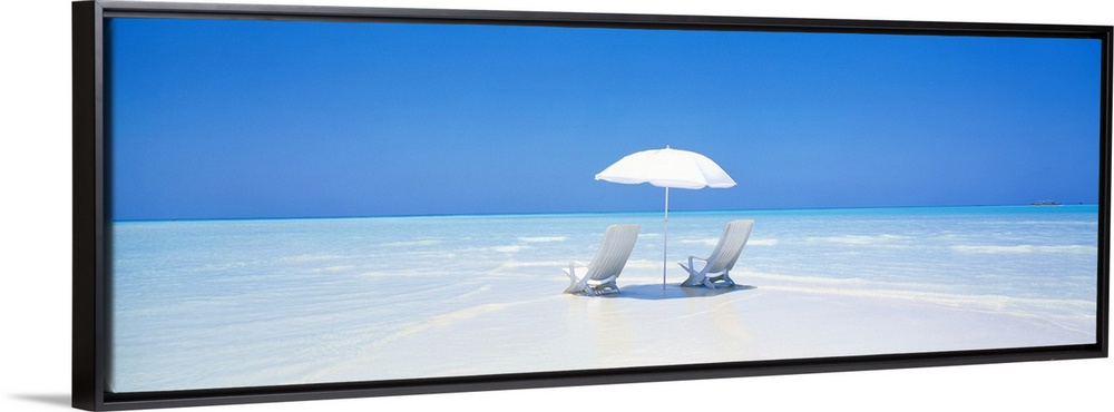 A panoramic photograph displaying two chairs sitting underneath an umbrella on a sandy beach in Maldives.  The clear water...