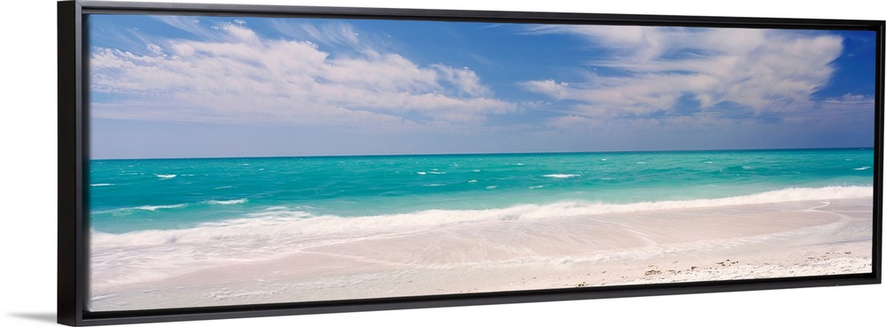 Panoramic photograph of calm ocean with surf and sand in the foreground and cloudy sky above.
