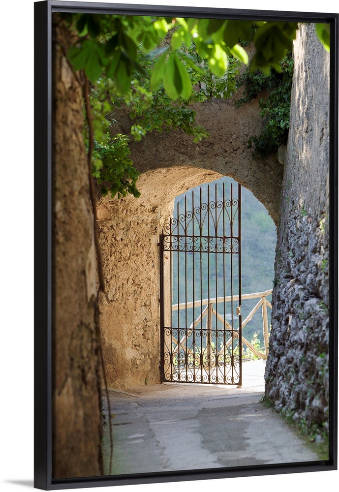 A vertical photograph of a walkway lined with old masonry a green vines to an open iron wrought gate.