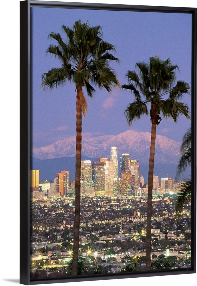 A vertical photograph of skyscrapers in the Los Angelesos skyline framed by two palm trees and mountains in the background.