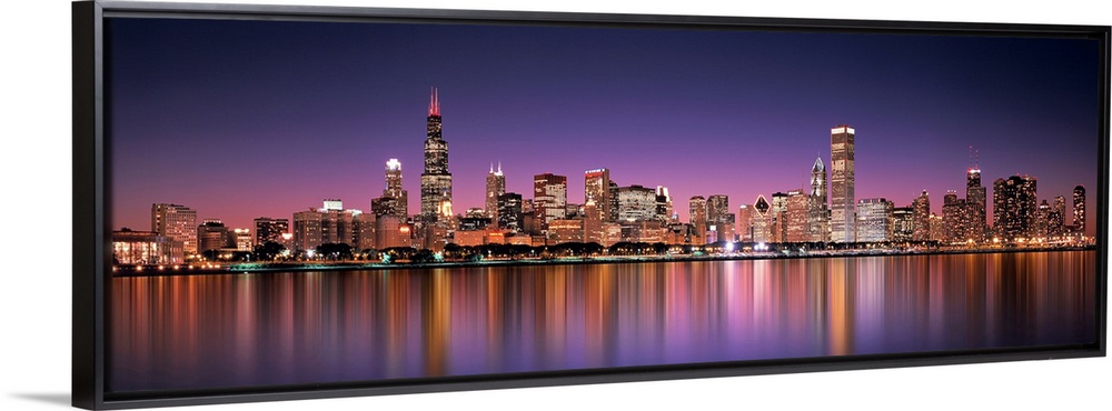 Panoramic photograph of the Chicago, Illinois skyline filled with skyscrapers reflecting onto Lake Michigan at night.