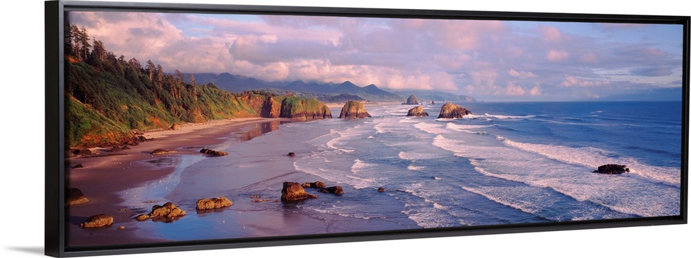 Panoramic photograph of waves slowly crashing against the rocks and sand of Cannon Beach in Oregon.  The mountains in the ...