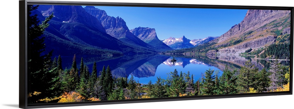 A photographic print of rugged mountains with snow in the distance reflected in a crystal clear lake with foliage in the f...