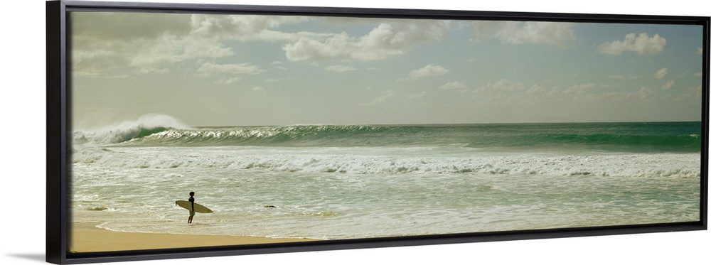 Panoramic image of a surfer standing where the ocean meets the beach shore looking at a big wave crashing.