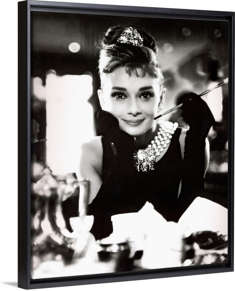 Classic iconic photograph of Audrey Hepburn sitting at a table holding a cigarettte and smiling.