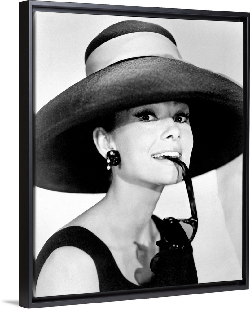 Large photograph of Audrey Hepburn in a giant sun hat holding sunglasses in her mouth as she glances at the camera in a bl...