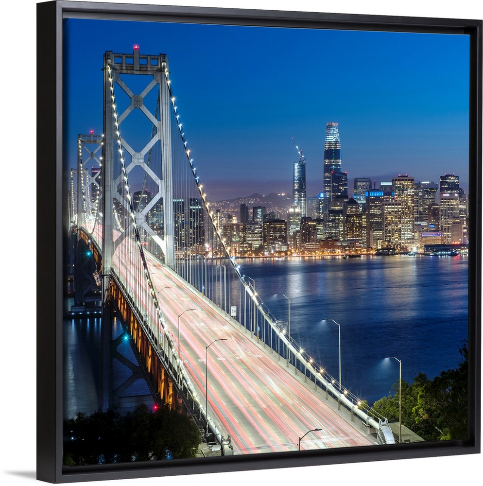 Square photograph of the Bay Bridge at dusk with downtown San Francisco lit up in the background.