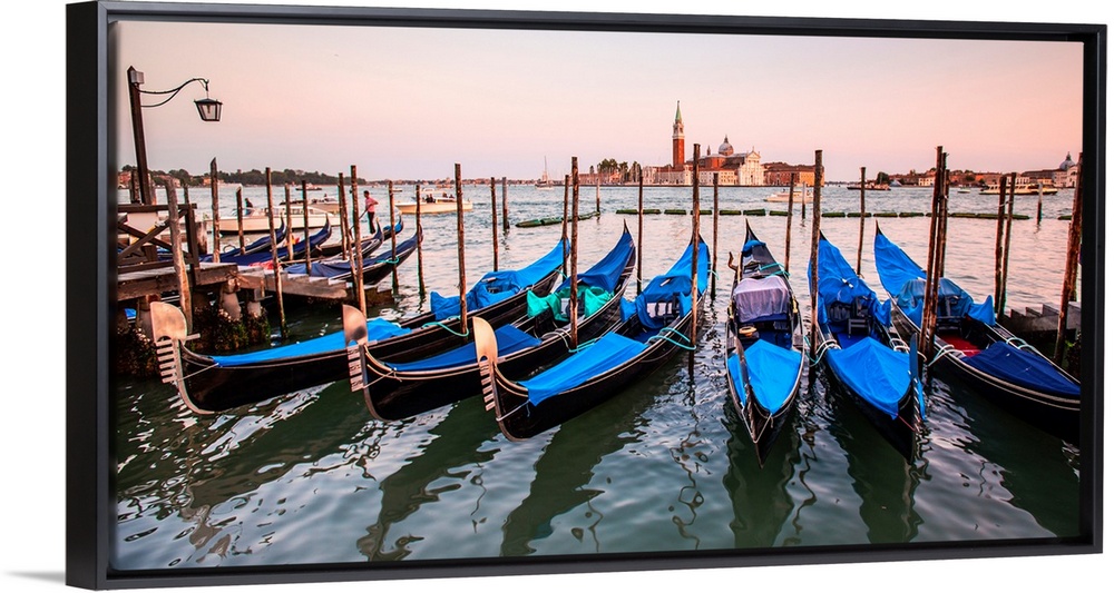 Panoramic photograph of blue gondolas docked in a row on the water with St. Mark's Square in the background at sunset.