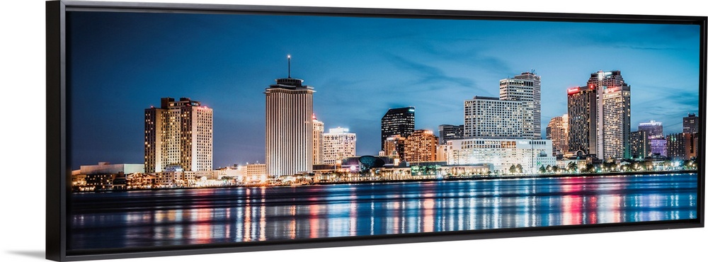 Panoramic photograph of the New Orleans skyline lit up at dusk and reflecting colorful bands onto the Mississippi River.