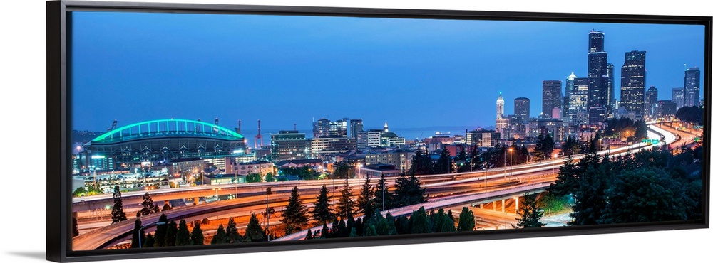 Panoramic photograph of the Seattle skyline with the stadium on the left and light trails from traffic on the highway.